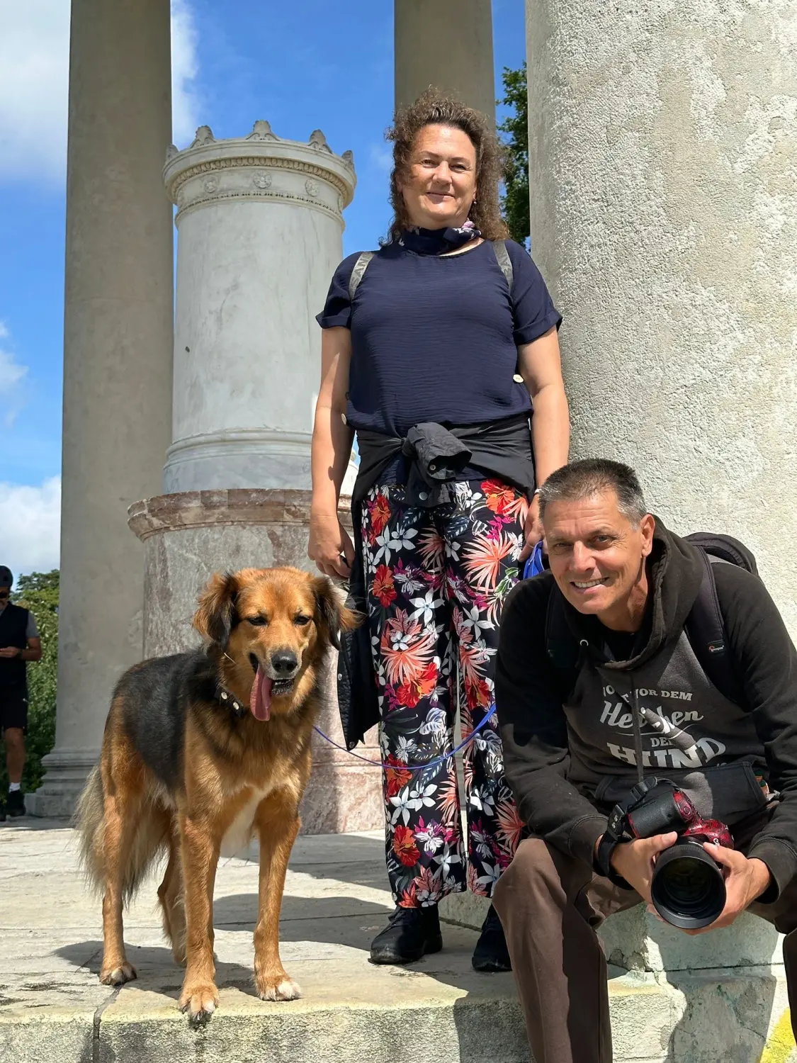 Paulchens-Hundeschule / Hunde-und Tierfotografie - Hundetraining und Fotografie - Ein Mann und eine Frau posieren mit einem großen Hund vor einer Steinsäule unter blauem Himmel.