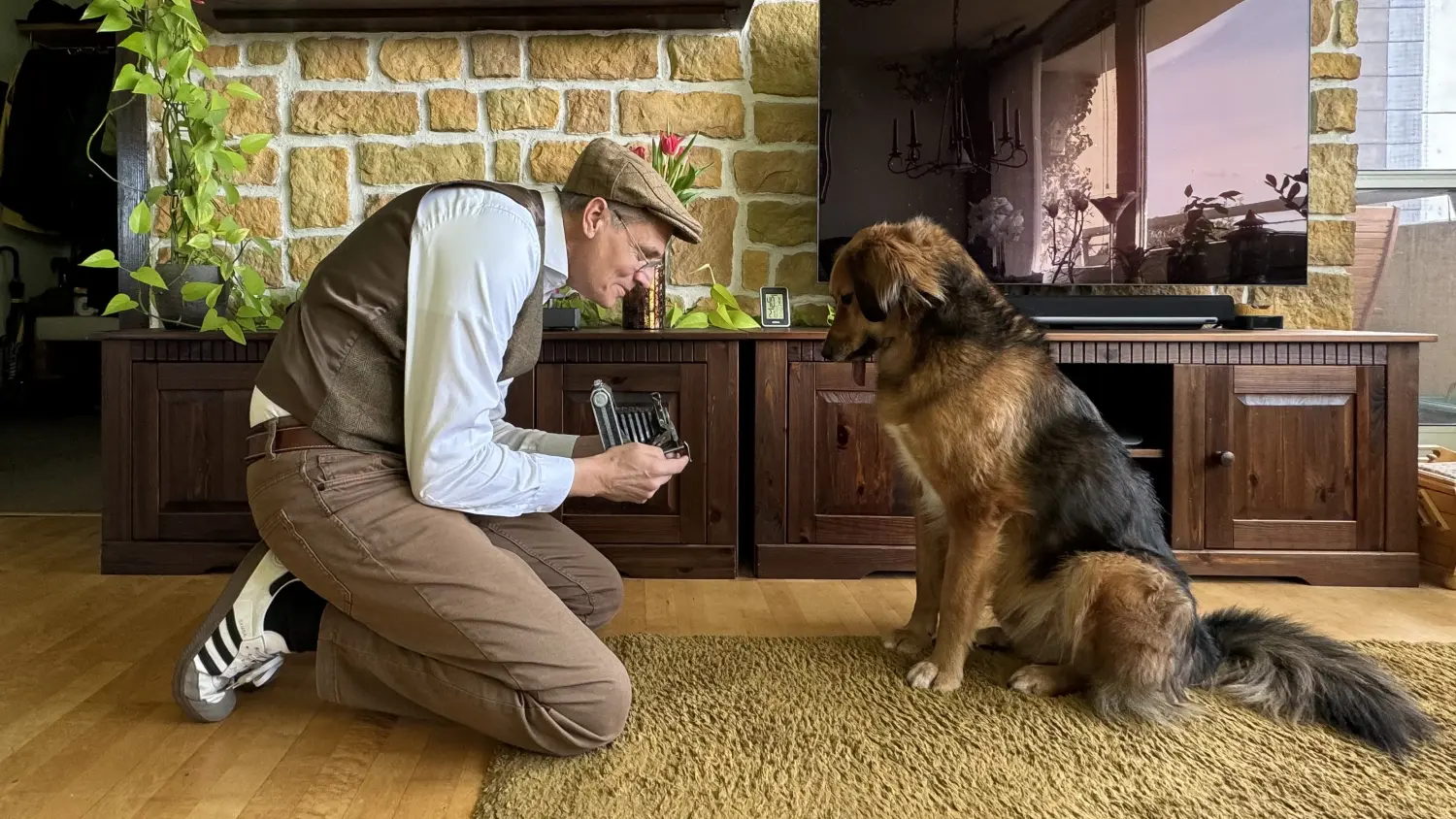 Paulchens-Hundeschule / Hunde-und Tierfotografie - Hundetraining und Fotografie - Ein Mann in Weste und Schiebermütze kniet drinnen auf einem Teppich und zeigt einem großen, langhaarigen braunen Hund einen kleinen Gegenstand.