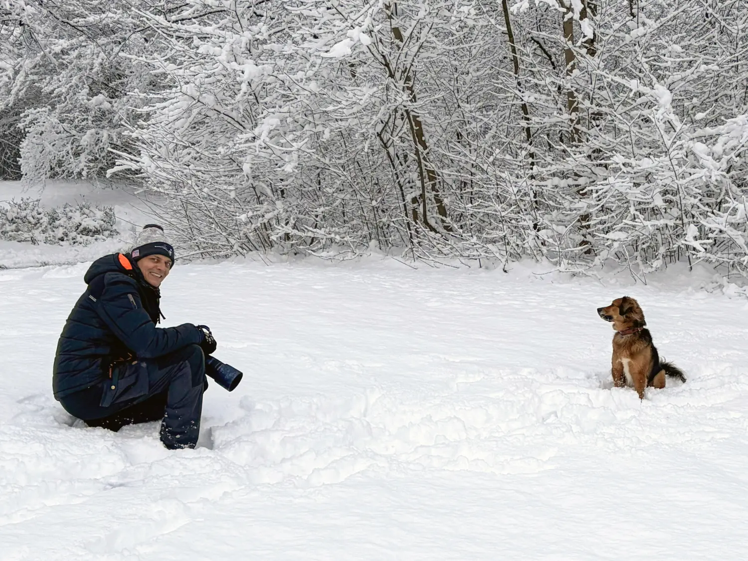 Paulchens-Hundeschule / Hunde-und Tierfotografie - Hundetraining und Fotografie - Der Mann kniet mit einer Kamera in der Hand und einem kleinen braunen Hund im tiefen Schnee.