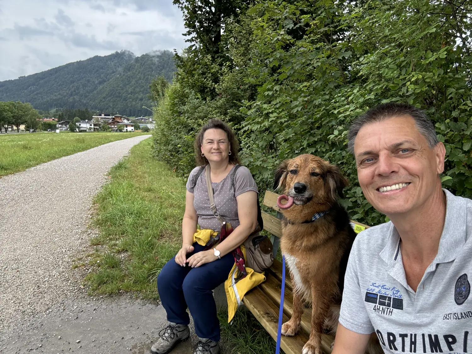 Paulchens-Hundeschule / Hunde-und Tierfotografie - Hundetraining und Fotografie - Eine Frau und ein Mann sitzen im Freien an einer üppig grünen Hecke. Zwischen ihnen sitzt ein mittelgroßer, brauner Hund, der spielerisch die Zunge herausstreckt.